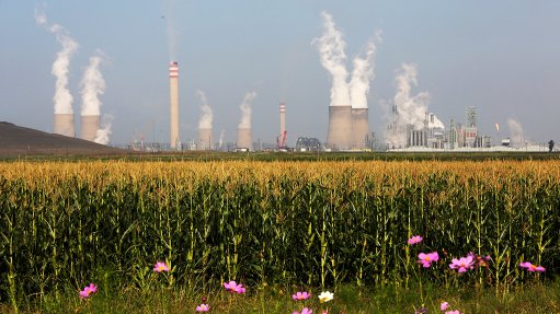 A photo of a Sasol plant emitting pollution into the air while crops and flowers grow in the foreground