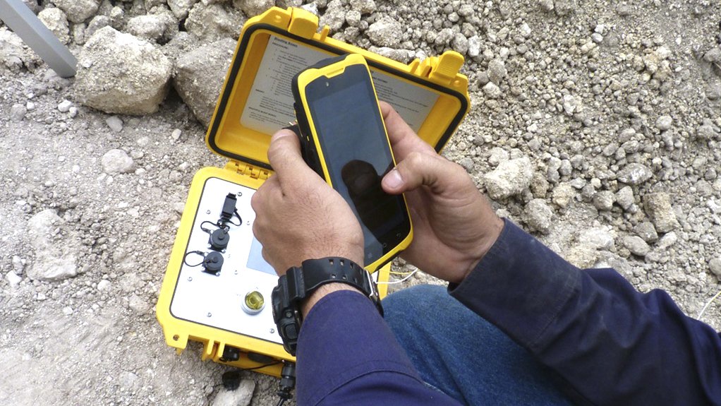 A yellow blasting mechanism linked to a cell phone being held in a hand, used to incorporate new technologies when blasting at mines