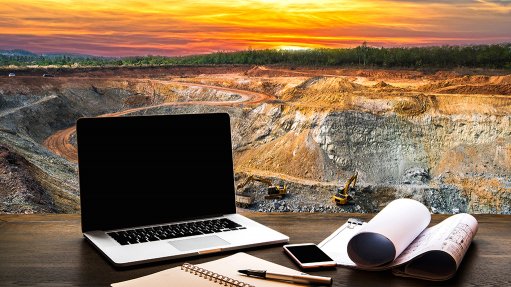 A sunset picture of a mine with yellow machines in operation, with a laptop, plans and cell phone in, while there is a green belt of natural vegetation in the background