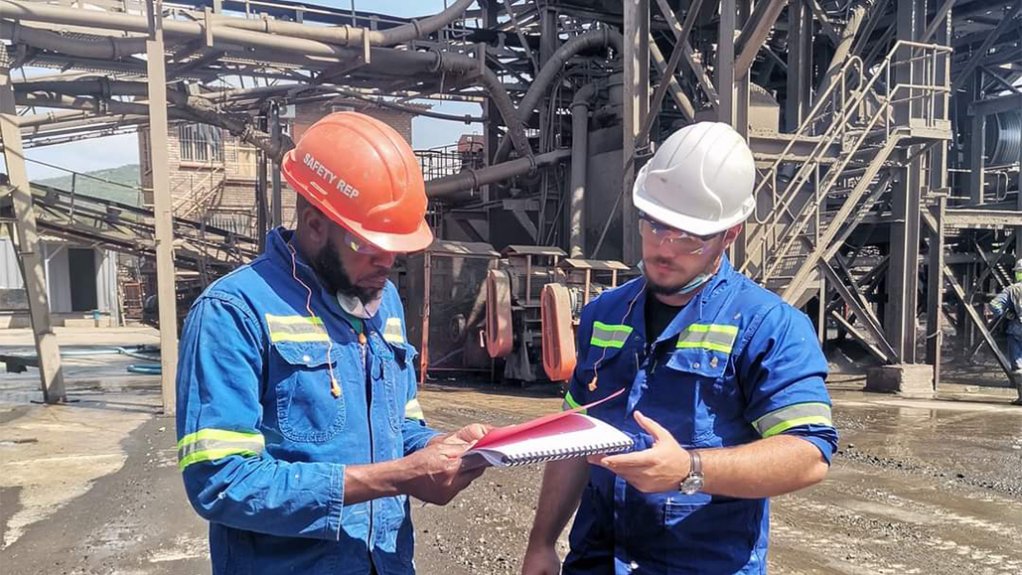 Two men in PPE in front of a mine's processing facility with a document which they are reading
