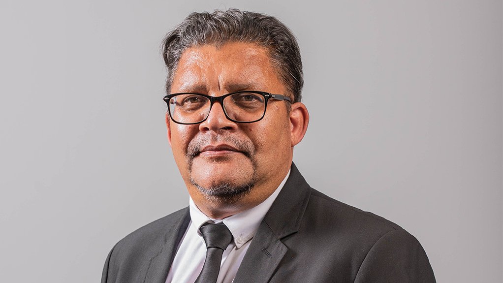 An image of Chris Campbell, standing with his hands in his pockets, wearing glasses, a black suit and tie with a white collared shirt