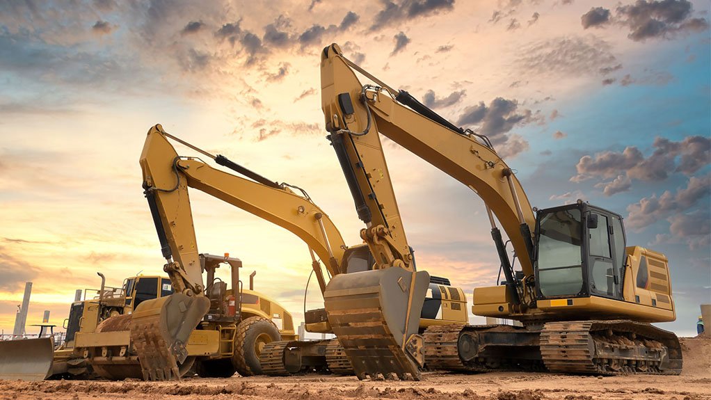 A row of capital equipment, or construction equipment, including graders and diggers on a construction site with the sun rise in the background