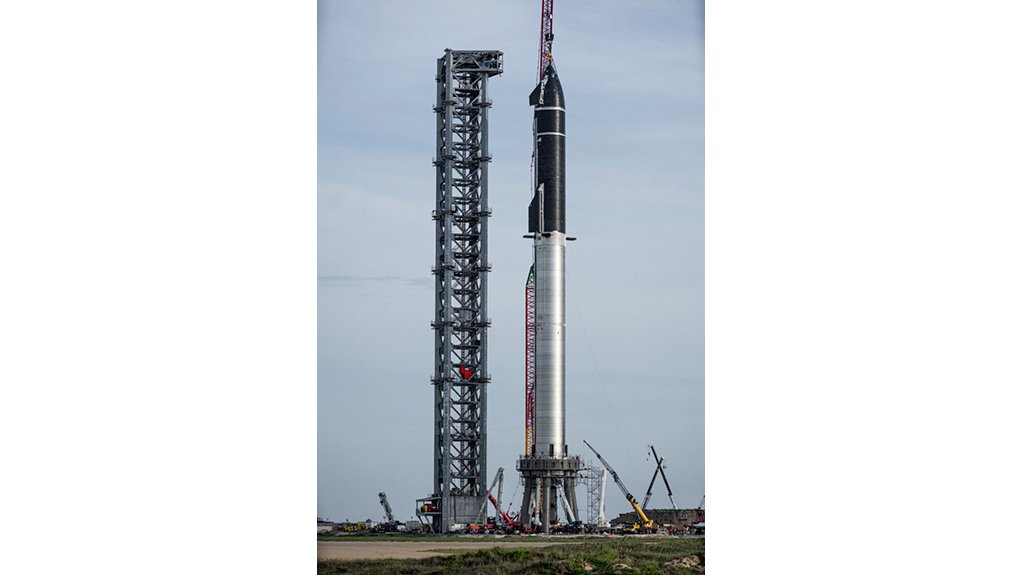 A Starship (covered in black thermal tiles) on top of a silver Super Heavy rocket, at Boca Chica