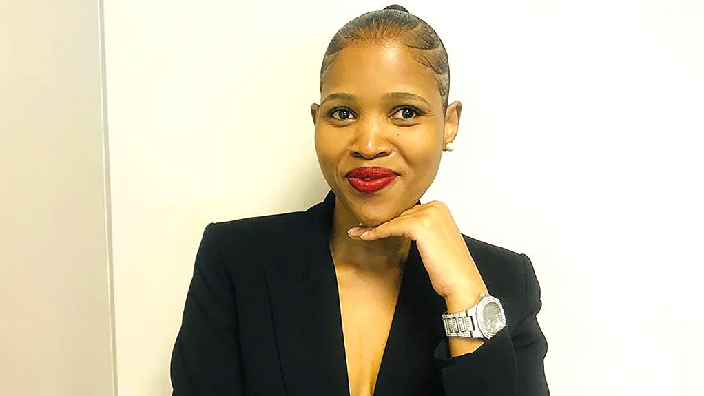 Lady in black blazer, braided up-style hairdo, smiling with hand under chin.