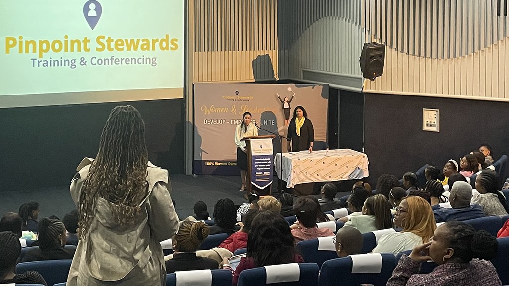 A group of ladies sat in a conference room with one at the foreground stood up asking the two hosting ladies a question.