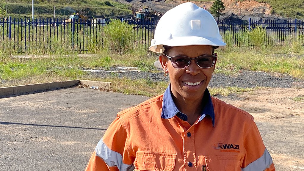 A photo of Maria Moganedi wearing PPE at the forefront of a mining operation with load haul vehicles in the background