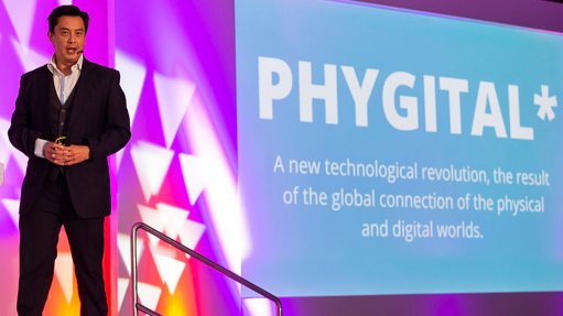 A gentleman giving a talk standing on a stage in front of a large screen with the word PHYGITAL