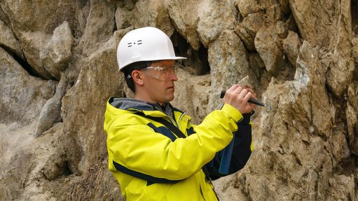 A man in yellow PPE in front of a rock face looking at a stone sample.