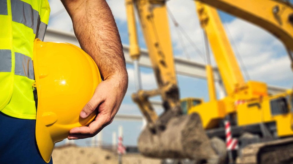 A man’s arm holding a hard hat wearing other PPE in the foreground of a construction site