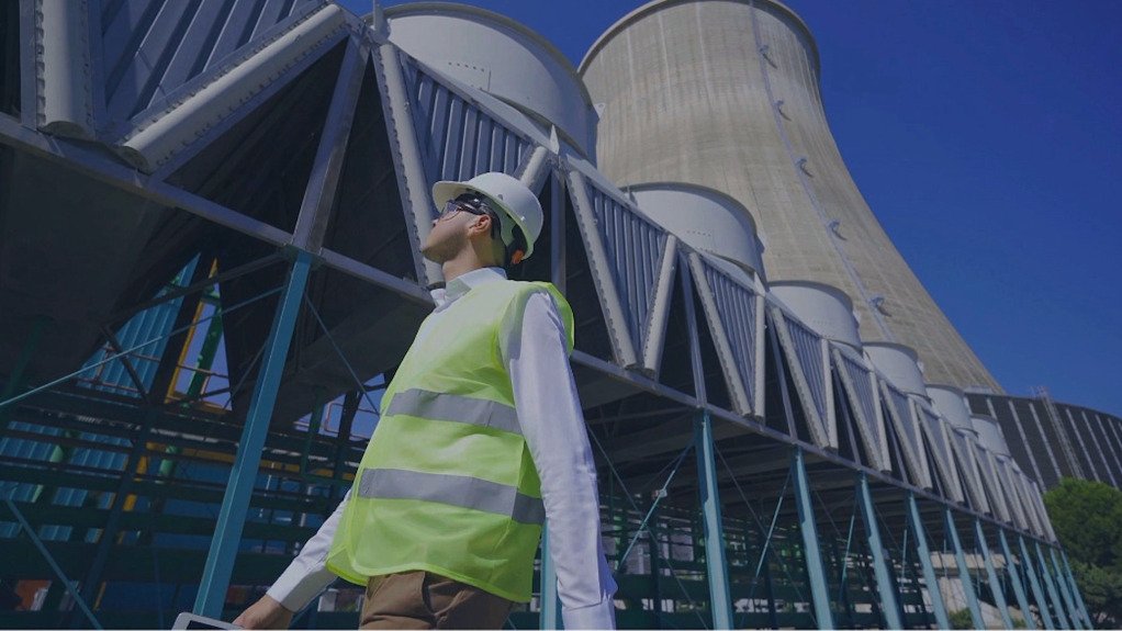 A man wearing PPE walking on front of Cooling towers looking up to the sky
