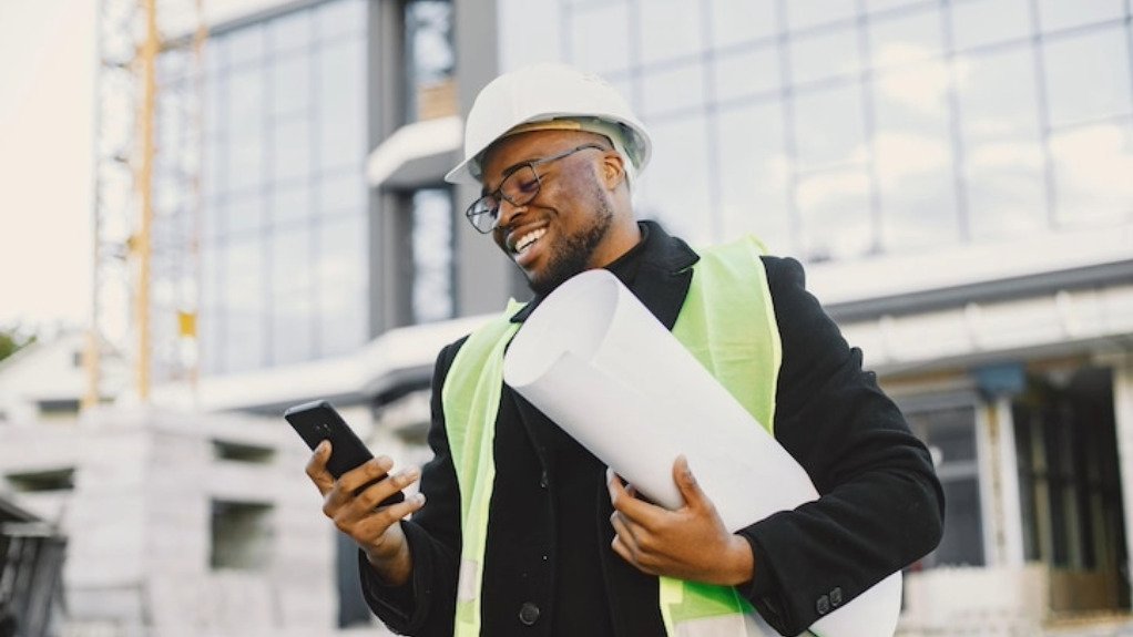 A young African man in a Visi vest holding business plans and a cell phone on front of a building