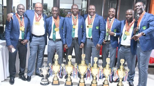 a group of rescuers and miners in suits in front of a row of trophies won at the 2025 International Mines Rescue Competition